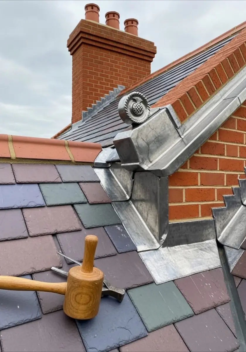 Metal flashing dressed where a slate roof meets the brickwork below a chimney, with a wooden mallet on the slates